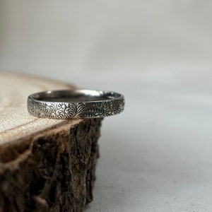 May include: A silver band ring with a delicate floral pattern engraved around the entire band.