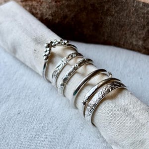 May include: A collection of silver rings with various designs, including beaded, etched, and smooth bands. The rings are displayed on a rolled white cloth, with a wooden background.