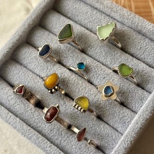 Puede incluir: Una colección de anillos hechos a mano con varios engastes de piedras preciosas, incluyendo vidrio marino y piedras de colores. Los anillos se exhiben en una bandeja de joyería de terciopelo gris. Los anillos tienen bandas de plata y formas de piedra únicas.