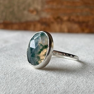 May include: A silver ring with an oval-cut gemstone, displaying green and white patterns. The ring has a hammered band and a bezel setting. The gemstone's intricate details are visible in this close-up image, showcasing its natural beauty.