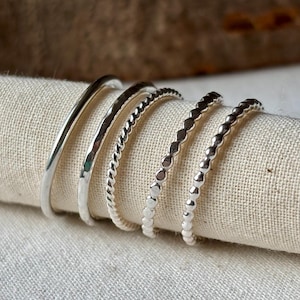 May include: Five silver rings of different designs are shown on a beige fabric. The rings include a smooth band, a hammered texture, a twisted rope pattern, and two beaded styles. The rings are suitable for stacking or wearing separately.