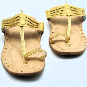 Könnte beinhalten: Ein Paar goldene Ledersandalen mit Zehenriemen und Knöchelriemen. Die Sandalen haben ein schlichtes Design und sind aus hochwertigem Leder gefertigt.