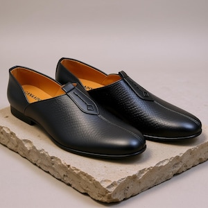 Puede incluir: Un par de zapatos negros de cuero sin cordones con un patrón texturizado. Los zapatos tienen un diseño cosido y un acento en forma de diamante en la pala. El interior es de color beige claro. Los zapatos se exhiben sobre una superficie de piedra.