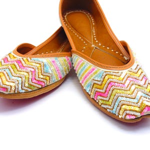 May include: A pair of white flat shoes with a zig-zag pattern of colorful sequins. The shoes have a brown leather insole and a white leather upper.