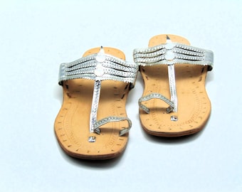 May include: A pair of silver leather sandals with a decorative strap across the top of the foot. The sandals have a brown leather sole and are made of a soft, supple leather.