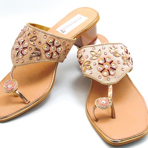 Pode incluir: Um par de sandálias de salto alto bege com detalhes dourados e embelezamentos florais intrincados. As sandálias têm um anel para o dedo do pé e uma parte de trás fechada.