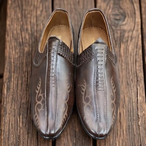May include: A pair of dark brown slip-on shoes featuring a smooth finish, a textured panel near the opening, and decorative laser-cut tribal patterns on the sides. The shoes have a central stitched seam running down the top and a tan interior lining.