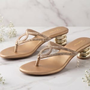 May include: A pair of gold-toned thong sandals featuring a sparkling rhinestone-encrusted strap design in an oval pattern. The sandals have a smooth footbed and a distinctive, stacked metallic gold cylindrical heel with a rounded shape.