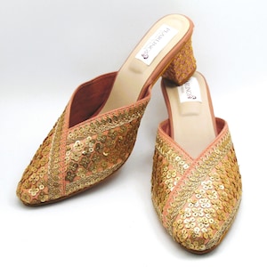 May include: A pair of peach-colored, open-back mules with gold sequin embellishments. The shoes have a low, block heel and a pointed toe. The brand name "PEARLINGS" is printed on the insole.