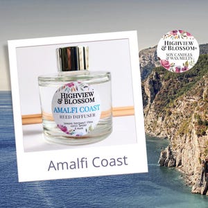 May include: A clear glass bottle with a silver lid and a white label that reads "Highview & Blossom Amalfi Coast Reed Diffuser Lemons, bergamot, Sitsea nerol amber & musk". The bottle is sitting on a wooden surface. The background is a blurry image of a rocky coastline and blue water.