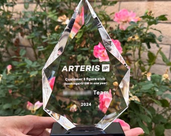 Op de afbeelding: Een heldere kristallen prijs met een zwarte basis. De prijs bevat de tekst "ARTERIS IP", "Customer 8 figure club (bookings>$10M in one year)", "Team" en "2024". De prijs wordt gehouden tegen een achtergrond van roze rozen en groene bladeren.