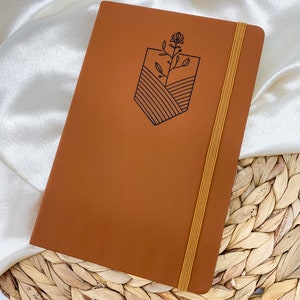 A brown leather bound journal with a yellow elastic closure. The journal has a simple line drawing of a flower and a landscape on the front cover.