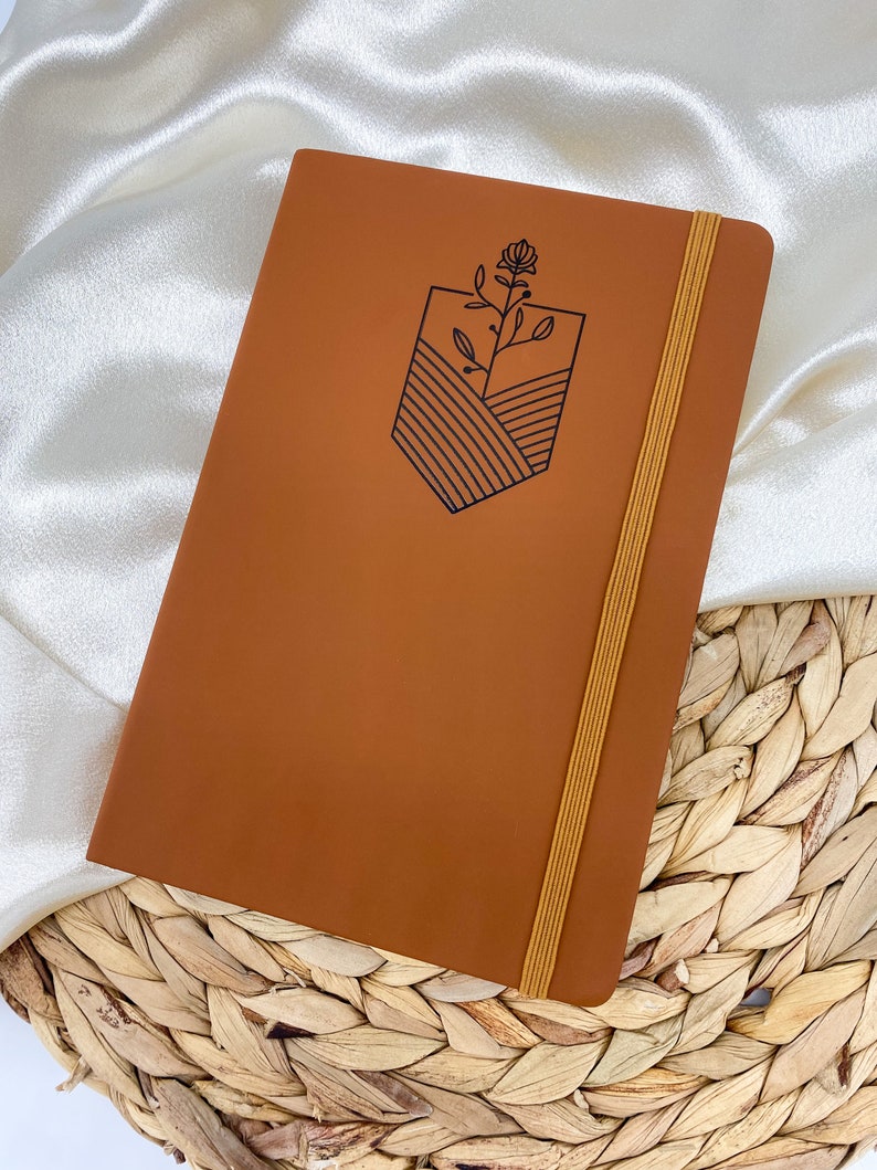 A brown leather bound journal with a yellow elastic closure. The journal has a simple line drawing of a flower and a landscape on the front cover.