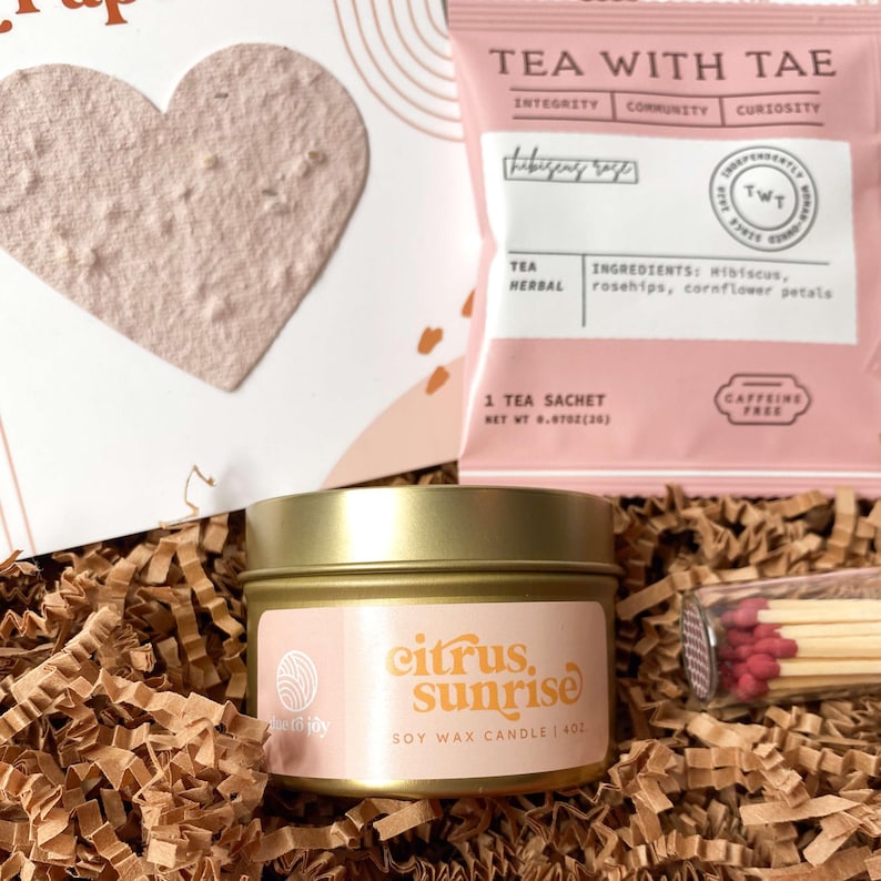 K&ouml;nnte beinhalten: Eine Geschenkbox mit einem herzf&ouml;rmigen Samenpapier, einem rosa Teebeutel mit der Aufschrift "Tea With Tae" und einer goldfarbenen Kerzendose mit dem Aufdruck "Citrus, Sunrise". Die Box enth&auml;lt auch eine kleine Schachtel Streichh&ouml;lzer.