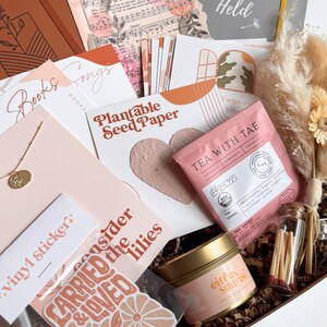 K&ouml;nnte beinhalten: Eine Geschenkbox mit verschiedenen Artikeln, darunter eine Halskette, Aufkleber, Tee, eine Kerze und Schreibwaren. Die Box enth&auml;lt eine "Plantable Seed Paper"-Karte, ein "Tea With Tae"-Teebeutel und eine "Citrus Sunrise"-Kerze. Die Farbgebung ist haupts&auml;chlich in Rosa, Orange und Braun gehalten.