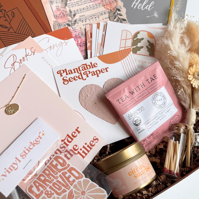 K&ouml;nnte beinhalten: Eine Geschenkbox mit verschiedenen Artikeln, darunter eine Halskette, Aufkleber, Tee, eine Kerze und Schreibwaren. Die Box enth&auml;lt eine "Plantable Seed Paper"-Karte, ein "Tea With Tae"-Teebeutel und eine "Citrus Sunrise"-Kerze. Die Farbgebung ist haupts&auml;chlich in Rosa, Orange und Braun gehalten.