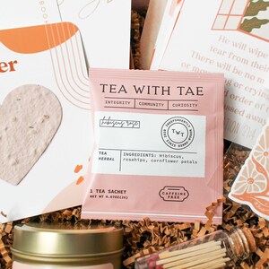 K&ouml;nnte beinhalten: Ein rosa Teebeutel mit der Aufschrift "TEA WITH TAE" und "hibiscus rose", umgeben von anderen Artikeln in einer Geschenkbox. Der Beutel listet die Zutaten auf: Hibiskus, Hagebutten und Kornblumenbl&uuml;ten. Eine Kerze und Streichh&ouml;lzer sind ebenfalls zu sehen.