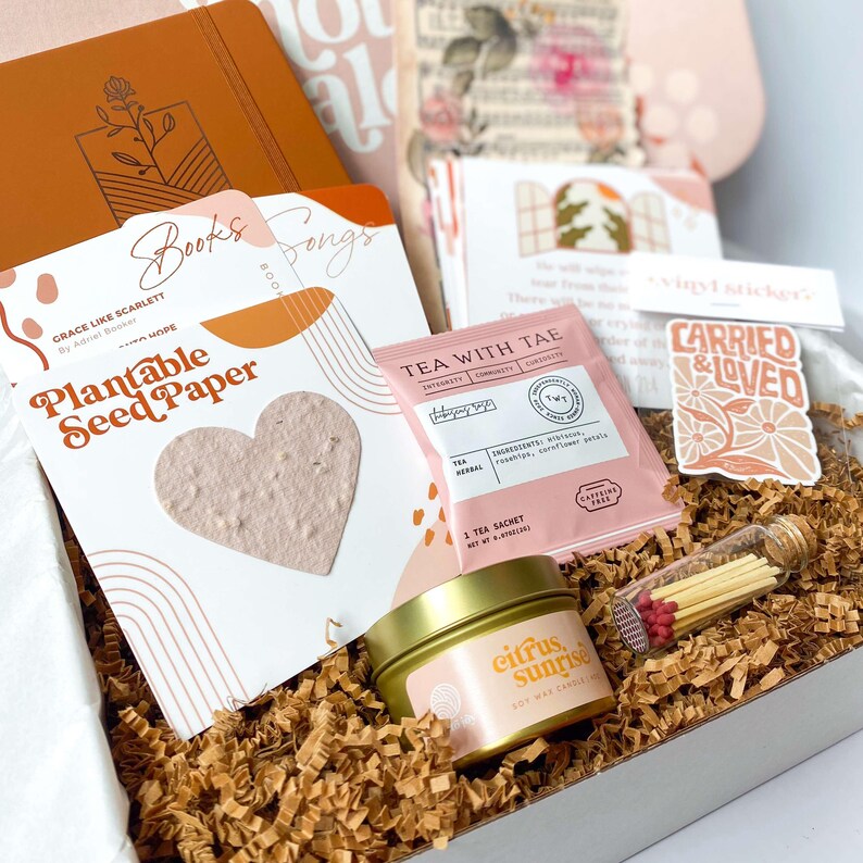 K&ouml;nnte beinhalten: Eine Geschenkbox mit verschiedenen Artikeln, darunter ein Notizbuch, Saatpapier, Tee, eine Kerze und Streichh&ouml;lzer. Das Saatpapier ist herzf&ouml;rmig. Die Kerze befindet sich in einer goldenen Dose und tr&auml;gt die Aufschrift "Citrus Sunrise". Ein Aufkleber lautet "Carried & Loved".