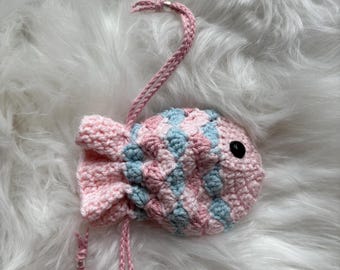 May include: A handmade crochet fish-shaped pouch in pink and light blue. The pouch has a drawstring closure and a black button eye. The fish is lying on a white, fluffy surface. The pouch is a unique and charming accessory.