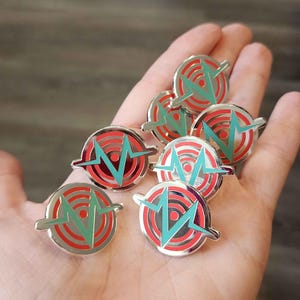 May include: A collection of eight silver pins with a red and teal design. The design features a stylized target with a lightning bolt in the center.