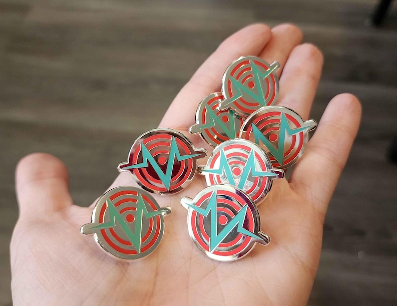 May include: A collection of eight silver pins with a red and teal design. The design features a stylized target with a lightning bolt in the center.