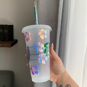 May include: A white reusable cup with a green straw and a holographic hibiscus flower design. The cup has the Starbucks logo on the side.