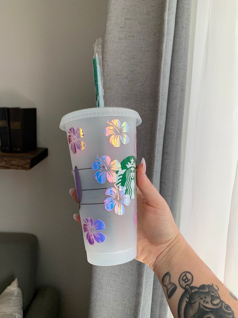May include: A white reusable cup with a green straw and a holographic hibiscus flower design. The cup has the Starbucks logo on the side.