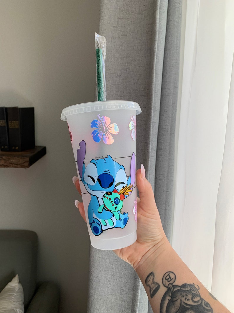 May include: A white reusable cup with a blue Stitch character holding a green doll. The cup has a clear plastic lid and a green straw. The cup is decorated with iridescent pink, blue, and purple flowers.