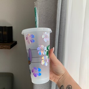 May include: A white reusable cup with a clear lid and a green straw. The cup has a holographic design of flowers and a cartoon character.