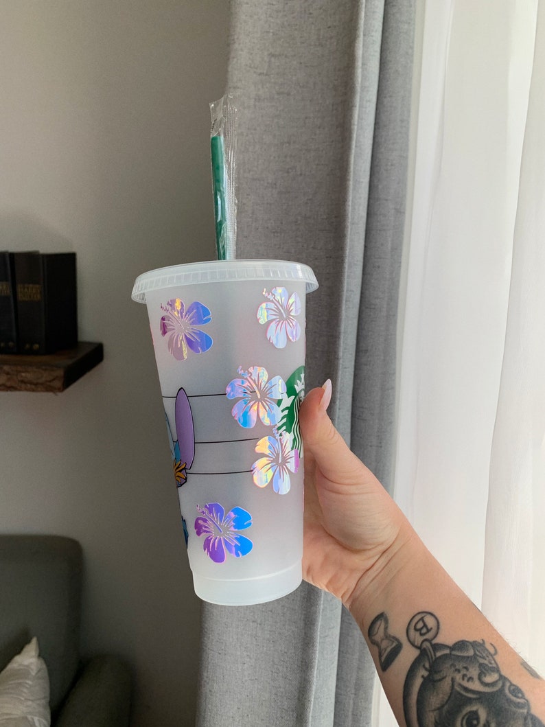May include: A white reusable cup with a clear lid and a green straw. The cup has a holographic design of flowers and a cartoon character.
