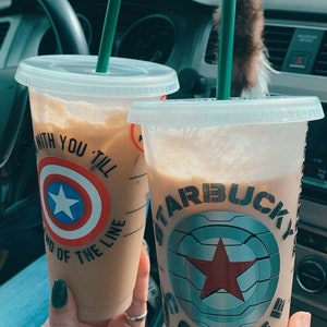 Op de afbeelding: Twee ijskoffie drankjes in doorzichtige plastic bekers met deksels en groene rietjes. De bekers hebben aangepaste ontwerpen met een rode ster en de tekst "STARBUCKS COFFEE" op de ene beker en een rood-wit-blauwe ster met de tekst "WITH YOU TILL END OF THE LINE" op de andere beker.