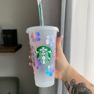 May include: A clear plastic reusable cup with a green Starbucks logo and iridescent pink, blue, and purple hibiscus flowers. The cup has a green straw.