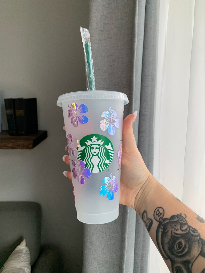 May include: A clear plastic reusable cup with a green Starbucks logo and iridescent pink, blue, and purple hibiscus flowers. The cup has a green straw.