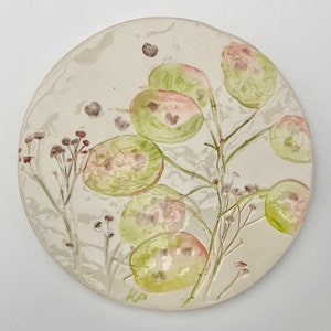 May include: A white ceramic plate with a delicate floral design. The design features green and pink flowers with a light brown stem. The plate is signed "KP" in the bottom right corner.