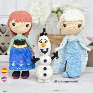 May include: Crocheted dolls of Elsa and Anna from the film Frozen, alongside Olaf the snowman. The dolls are dressed in their signature outfits, with Elsa in a blue gown and Anna in a blue and green dress. The dolls are standing on a white surface with a white background.