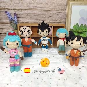 Peut inclure: Une collection de cinq personnages de Dragon Ball Z en crochet, dont Goku, Vegeta, Bulma, Gohan et Krilin. Les personnages sont debout sur une surface blanche avec une boule de dragon jaune au centre. L'arrière-plan est une étagère en bois avec un cadre photo et une plante en pot.