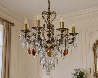 May include: An ornate brass chandelier featuring six candle-style light sockets, intricate metal scrollwork, and numerous hanging clear crystal prisms. Several amber and deep red teardrop-shaped crystals are interspersed among the clear ones.