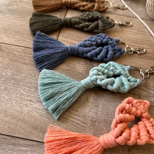 May include: A set of seven colorful macrame keychains with tassels. The keychains are in the shape of a heart and are made with a variety of colors, including white, brown, green, blue, light blue, and orange.