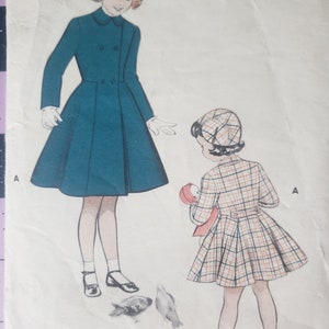 May include: A vintage sewing pattern featuring two girls wearing coats. The girl on the left is wearing a teal coat with a white collar and white gloves. The girl on the right is wearing a plaid coat with a red and white hat. Both girls are wearing black shoes. The pattern is labeled "A" on both sides.