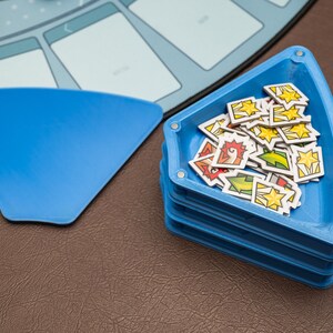 May include: A stack of blue, triangular game piece trays with magnets, filled with colorful game tokens. A matching blue game piece is next to the trays. The game pieces have various designs, including stars and other symbols. The game is on a brown surface.