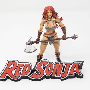 Może przedstawiać: Figurka Red Sonji z rudymi włosami, w srebrnym i brązowym stroju, trzymająca srebrny topór i broń z kolcami. Figurka stoi przed czarno-czerwonym logo "Red Sonja".