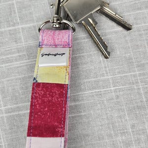 Könnte beinhalten: Ein farbenfrohes Stoff-Schlüsselband mit einem silbernen Karabiner und Schlüsselring. Das Schlüsselband hat ein Patchwork-Design in Rosa, Gelb und Rot. Ein kleines weißes Etikett mit schwarzem Text ist auf das Schlüsselband genäht. Zwei Schlüssel sind befestigt.