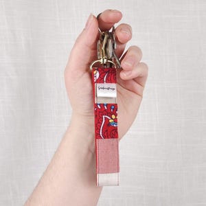 Könnte beinhalten: Ein rotes Schlüsselband aus Stoff mit Muster und goldfarbener Hardware. Das Schlüsselband hat ein Blumenmuster und den Text "SeafarerDesign". Das Schlüsselband wird in einer Hand vor weißem Hintergrund gehalten.