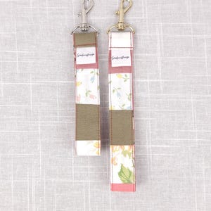 Könnte beinhalten: Zwei Schlüsselanhänger aus Stoff mit Metallverschlüssen. Die Schlüsselanhänger haben ein Patchwork-Design mit floralen und einfarbigen Blöcken in Grün, Rosa und Weiß. Die Verschlüsse sind silber- und goldfarben.