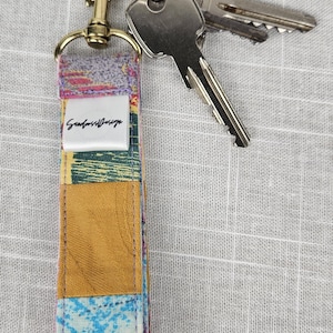 Könnte beinhalten: Ein farbenfrohes Stoff-Schlüsselband mit einem goldfarbenen Verschluss und Ring, an dem zwei Schlüssel befestigt sind. Das Schlüsselband hat ein Patchwork-Design mit verschiedenen Farben, darunter Blau, Gelb und Lila. Der Text "Seabreeze Design" ist sichtbar.