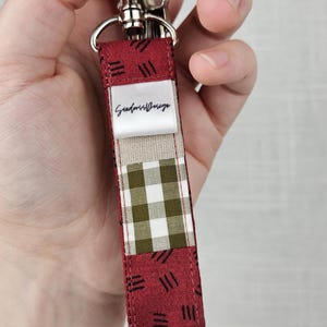 Könnte beinhalten: Ein Schlüsselanhänger-Band mit silbernem Verschluss und Ring. Das Band hat ein Patchwork-Design mit bordeauxfarbenem Stoff und einem grün-weiß karierten Muster. Der Text "Goddess Design" ist auf einem beigen Stoff aufgedruckt.