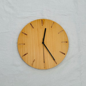 Könnte beinhalten: Eine runde Holzuhr mit schwarzen Zeigern und einer hellen Holzmaserung. Das Zifferblatt der Uhr hat ein schlichtes Design ohne Zahlen, nur Stundenmarkierungen.