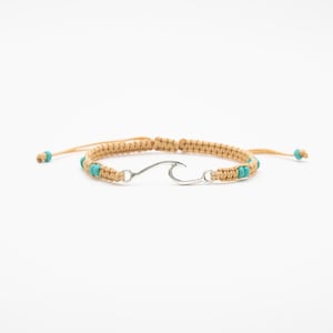 May include: A beige macrame cord bracelet featuring a central silver-toned metal wave charm. The bracelet is accented with small, round, turquoise-colored beads integrated into the woven cord design, which includes adjustable pull-string ends.