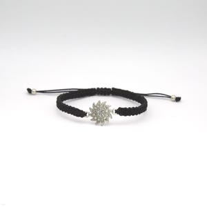 May include: A black cord bracelet featuring a central silver-toned sun-shaped charm adorned with small clear crystals. The bracelet has an adjustable macrame-style woven band with two small silver-toned beads at the ends of the pull strings.
