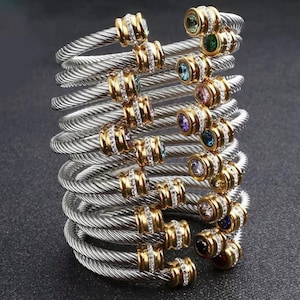 May include: A stack of silver-toned cable bracelets featuring twisted metal bands. Each bracelet is accented with gold-toned end caps embedded with small clear stones and a single round, multi-colored gemstone in various hues like green, blue, and pink.
